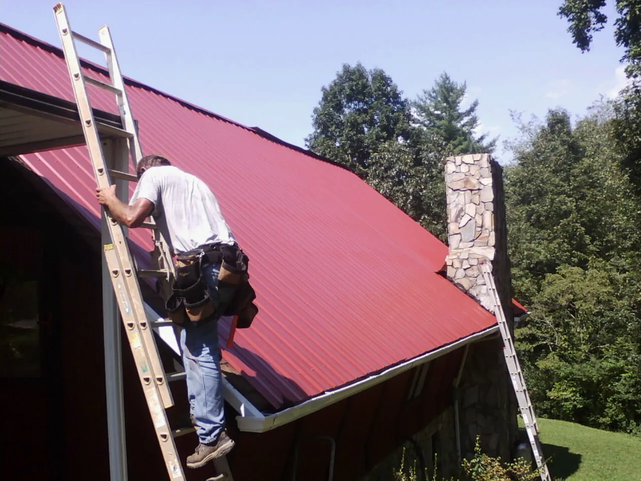 Roofer on a ladder beside a red metal roof and stone chimney — Soffit & Fascia Repair by Roofing Brothers in Rives