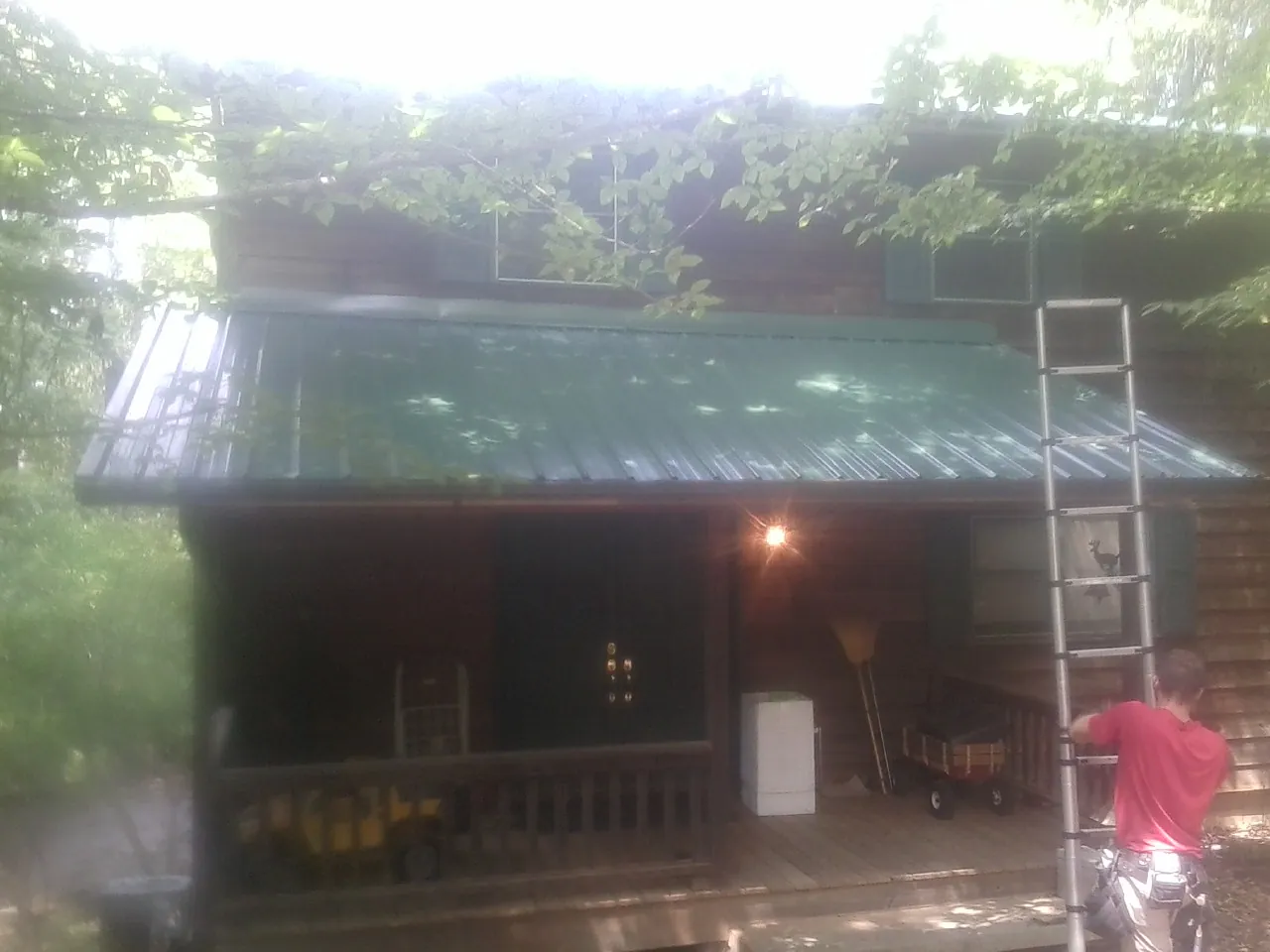 Worker on a ladder at the green porch roof of a wooded cabin in Rives